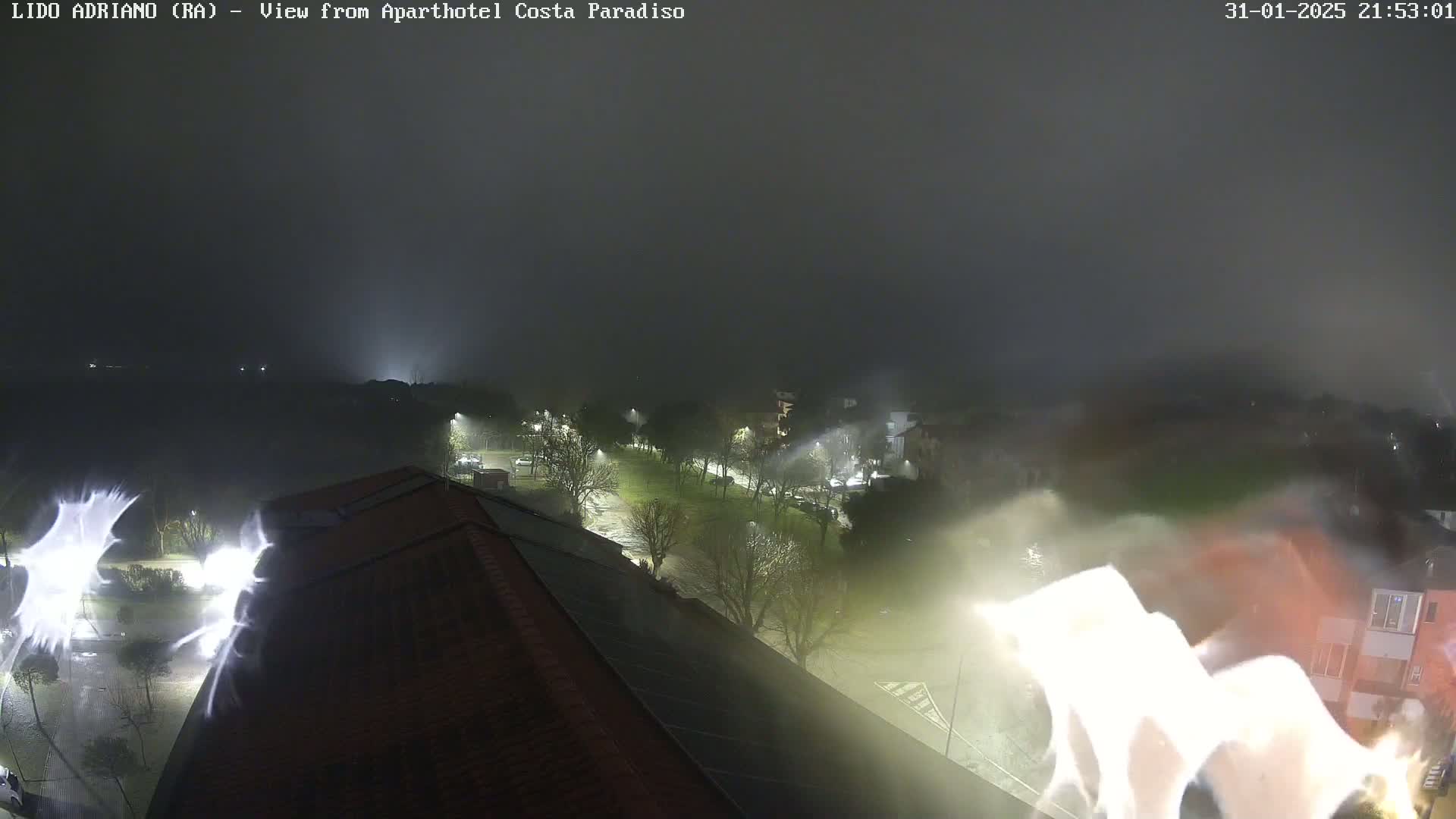 A nighttime, foggy view of a street and buildings, illuminated by streetlights, with bright light streaks possibly from lightning.