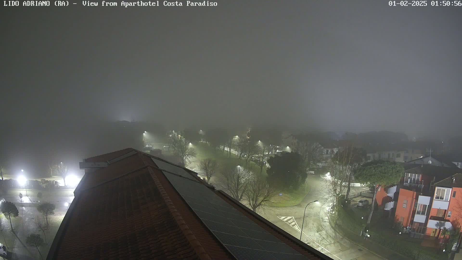 A foggy night scene shows a residential area with dimly lit streets and buildings, viewed from a rooftop with solar panels.