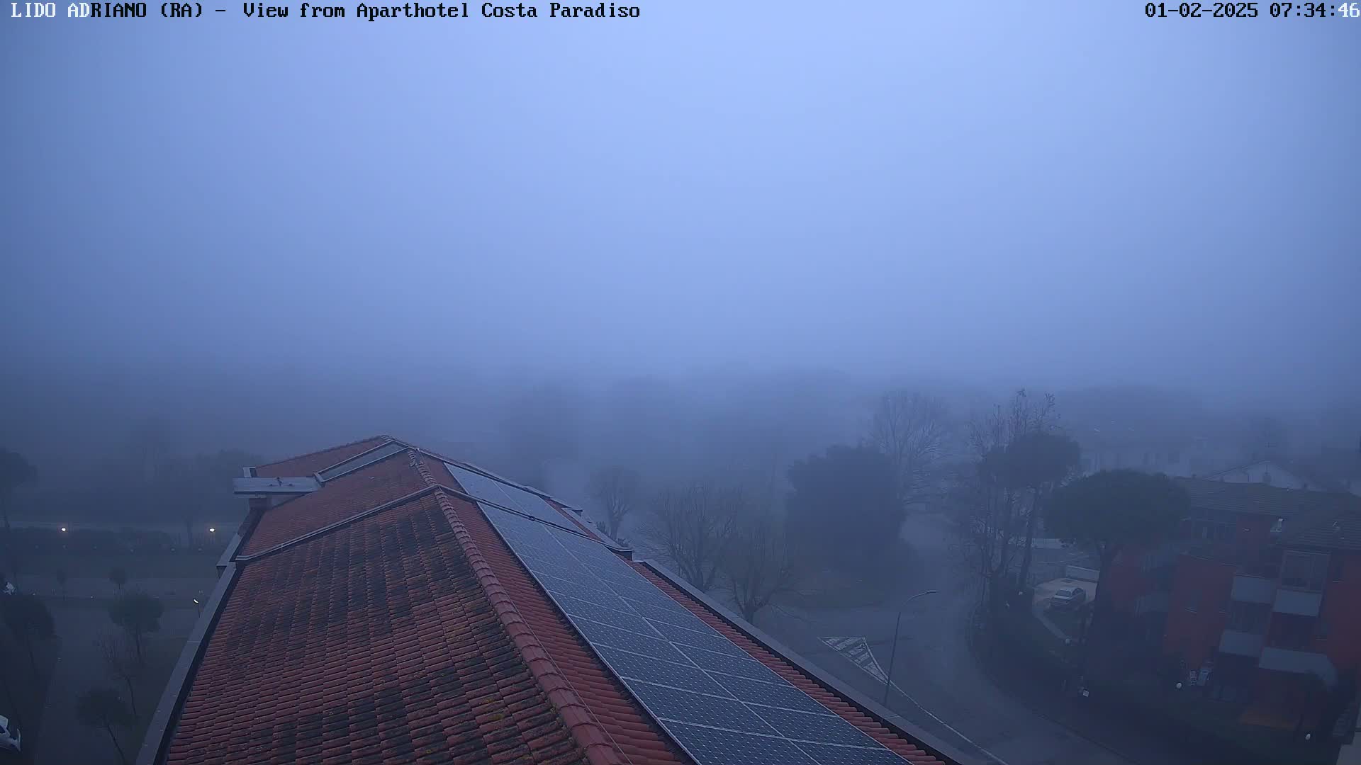 A thick fog obscures a view of a building's red tiled roof with solar panels, overlooking a street and other buildings in the distance.