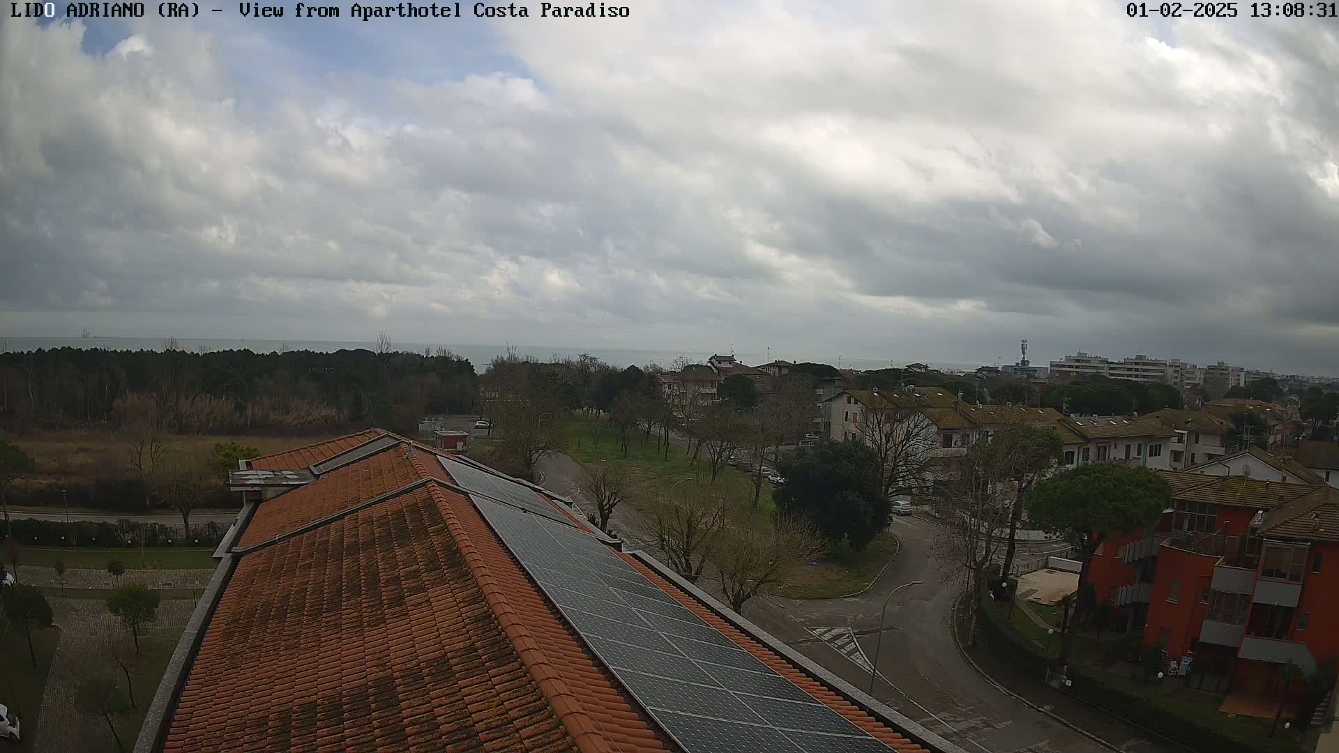 A view from a rooftop with solar panels shows a residential area under a cloudy sky, with a calm sea visible in the distance.