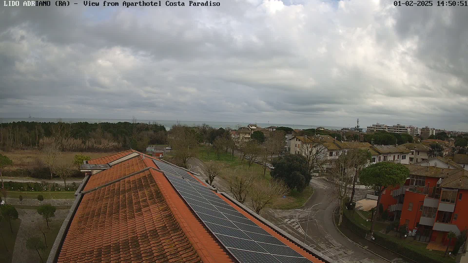 A view from a rooftop with solar panels shows a cloudy day overlooking a coastal town with residential buildings and a calm sea in the distance.