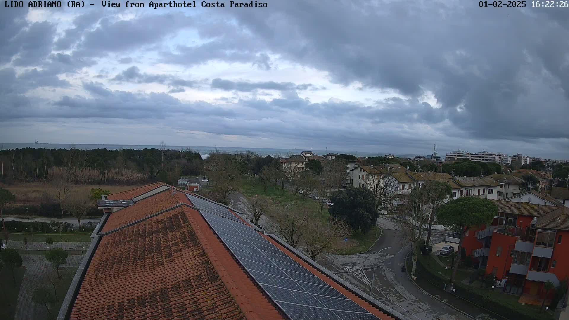 A cloudy day overlooks a coastal town with residential buildings, trees, and a partially visible sea in the distance, viewed from a rooftop with solar panels.