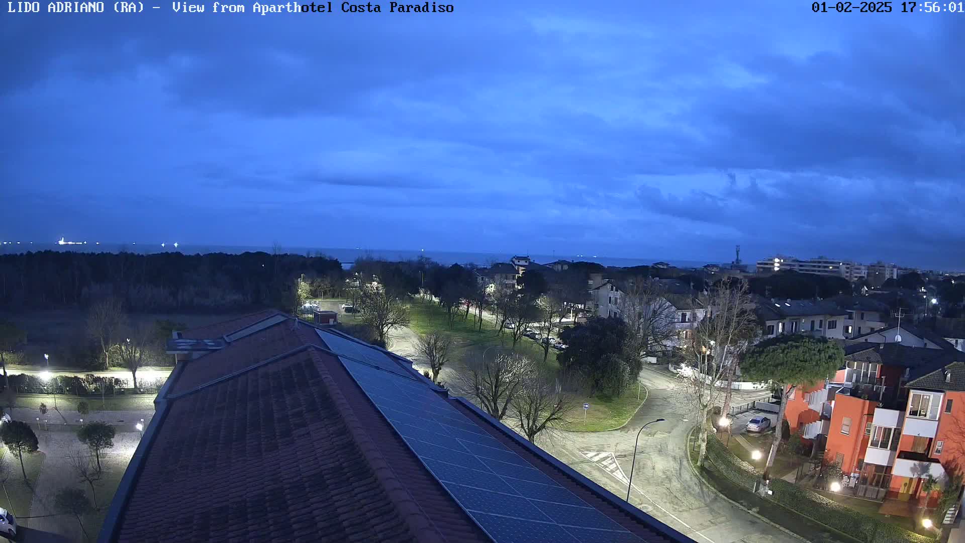 A twilight view of a coastal town shows a residential area with a park, roads, and buildings, including a building with solar panels on its roof, under a mostly cloudy sky.