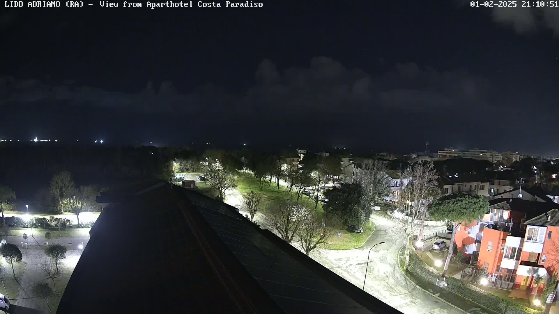 A nighttime, high-angle view of a residential area with streets, trees, and buildings, lightly clouded skies.