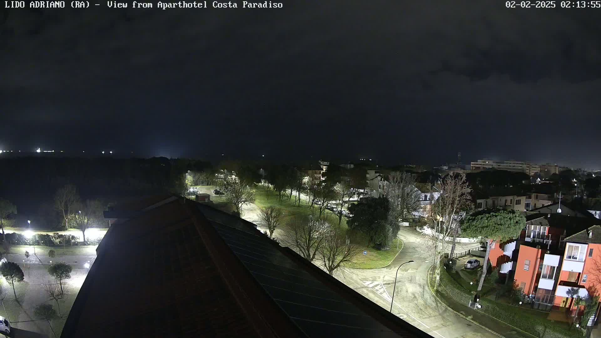 A nighttime view of a residential area with streetlights illuminating wet roads and buildings under a dark, cloudy sky.