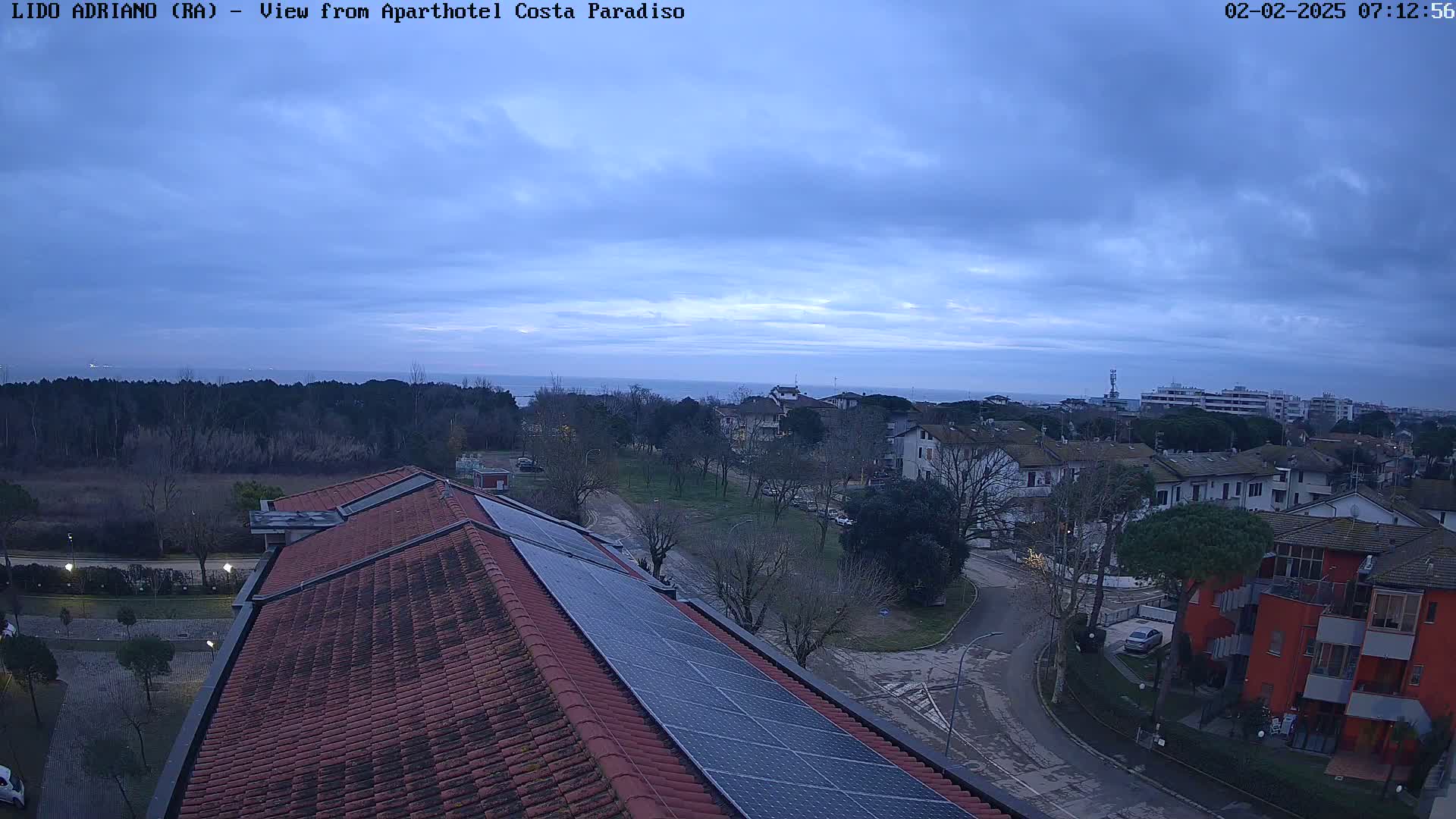 A rooftop with solar panels overlooks a residential area and a calm sea under an overcast sky.