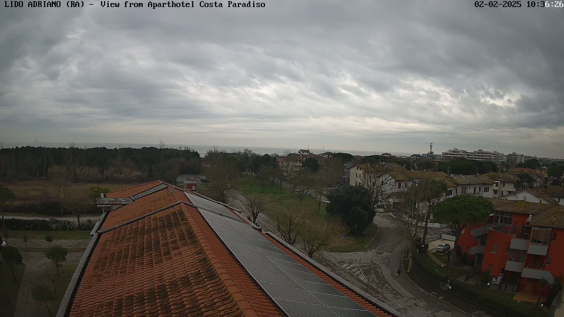 An overcast sky over a coastal town viewed from a rooftop with solar panels, showing a residential area with trees, roads, and a glimpse of the sea in the distance.