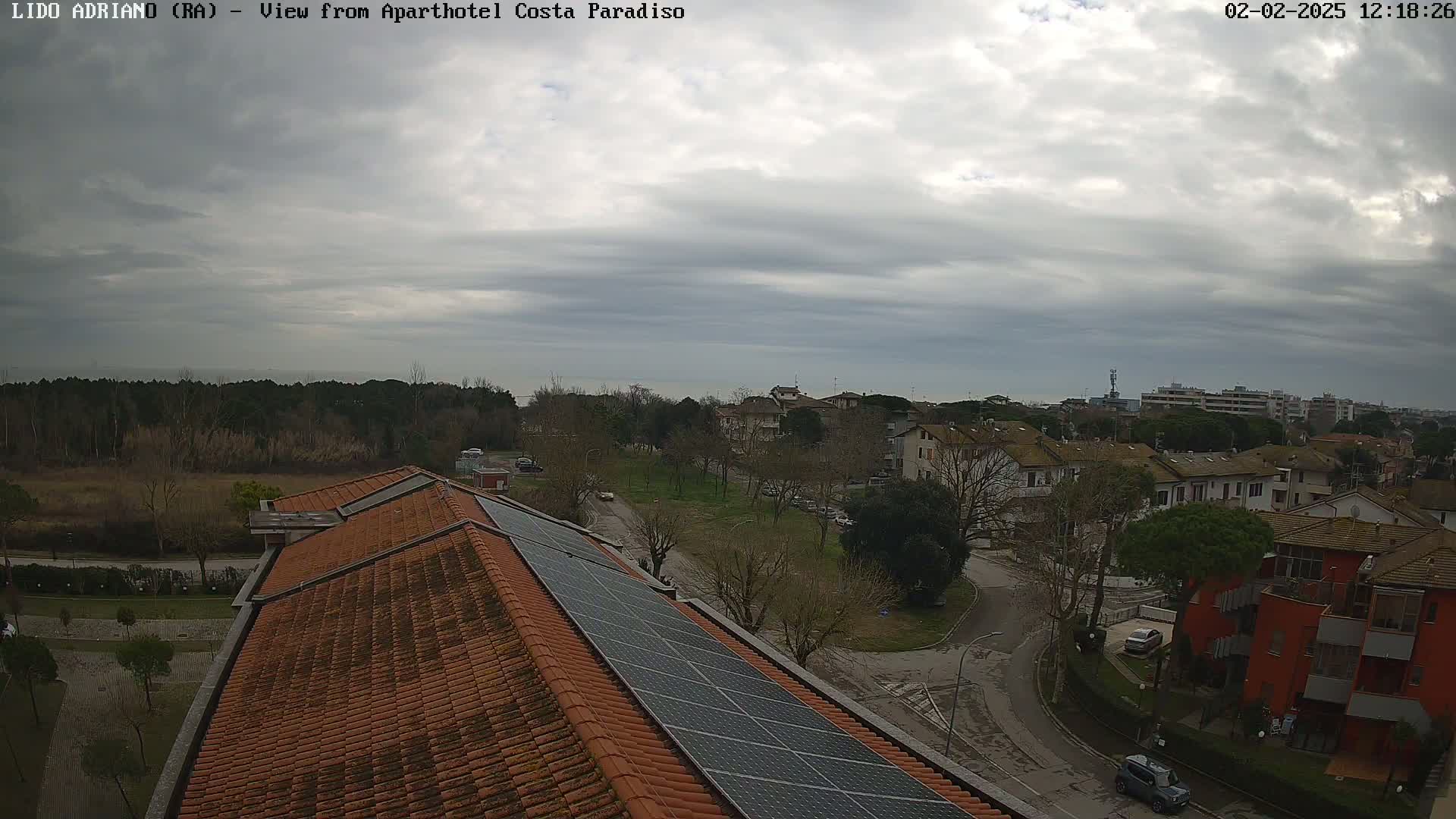 A mostly cloudy day overlooks a residential area with a rooftop featuring solar panels, a distant sea, and bare trees.