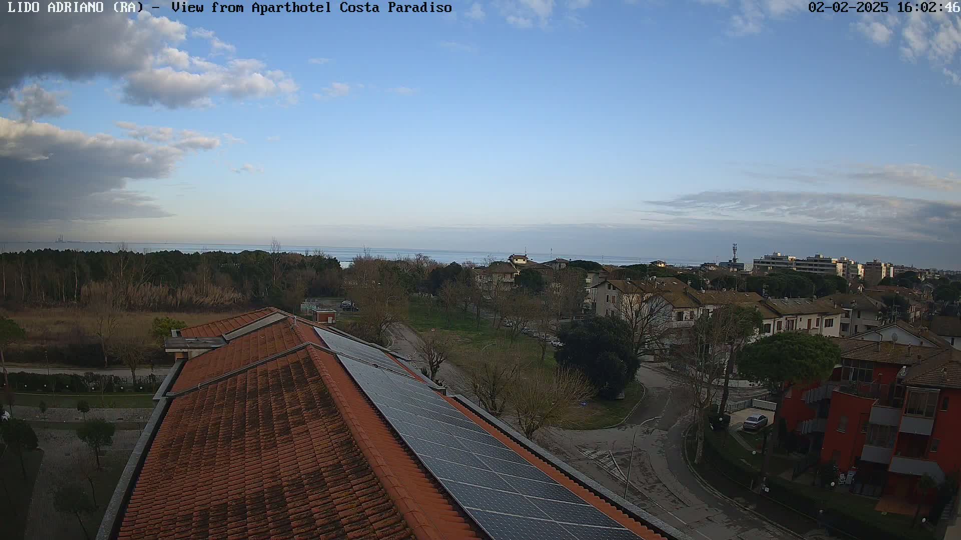 A partly cloudy sky overlooks a residential area with a calm sea in the distance, featuring a building with solar panels on its roof.