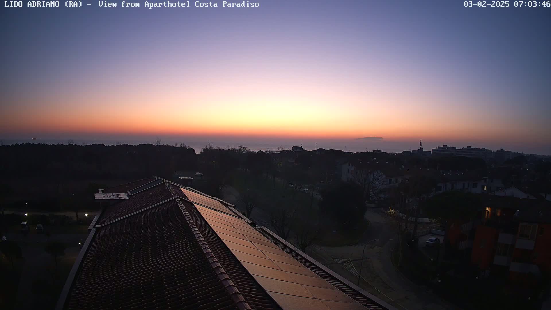 A rooftop with solar panels overlooks a coastal town at sunrise, with a calm sea visible in the distance.