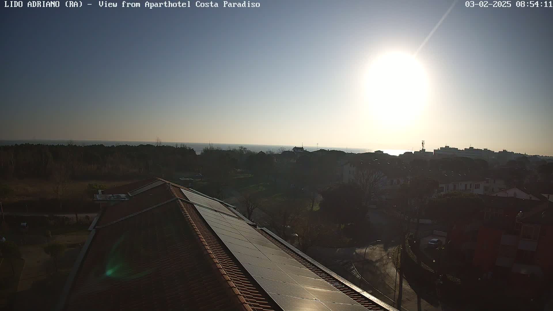 An aerial view shows a building with solar panels on its roof, overlooking a town next to a calm sea under a bright, sunny sky.