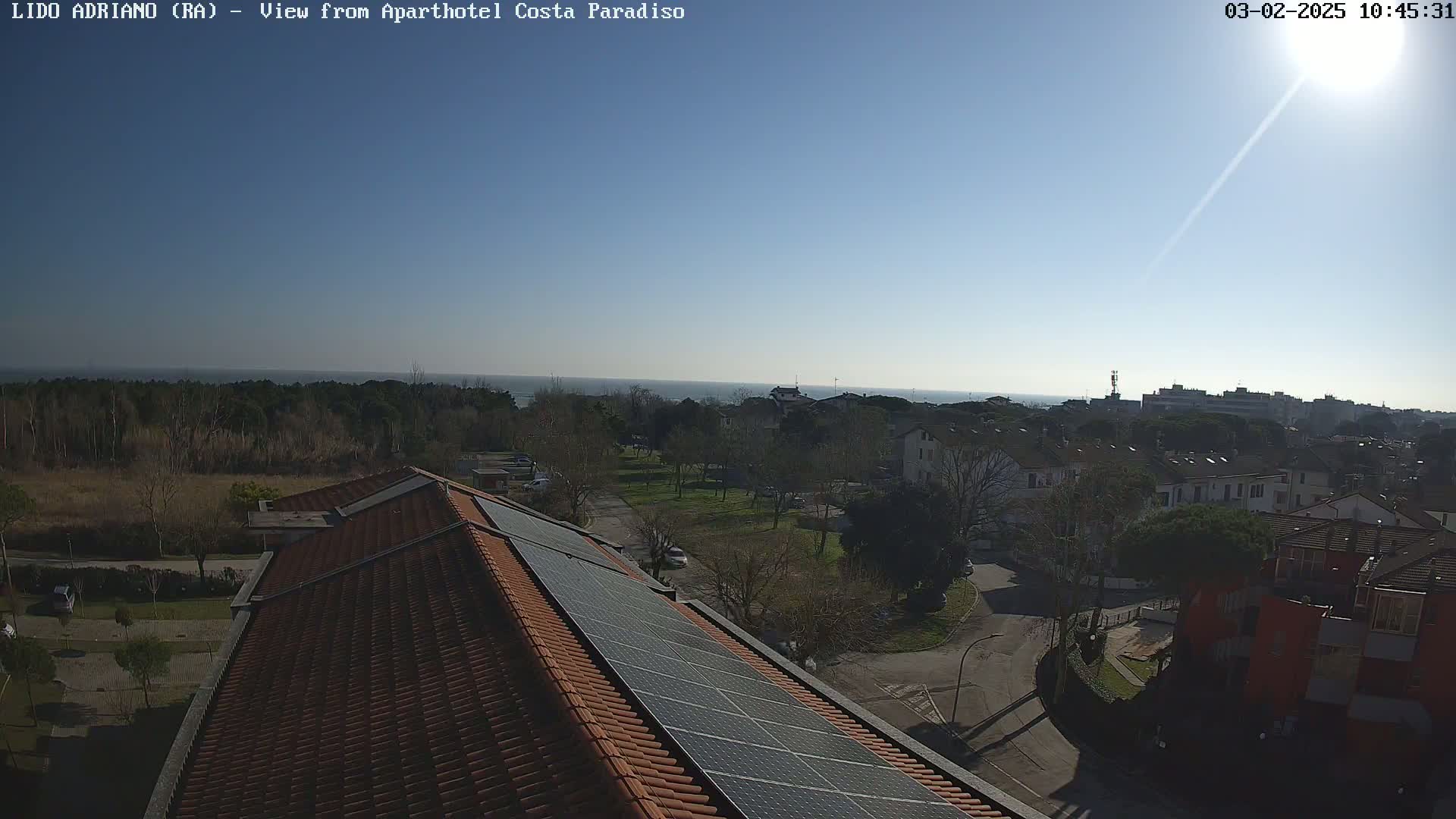 A sunny day reveals a coastal town's rooftops, a strip of trees, and a calm sea in the distance.