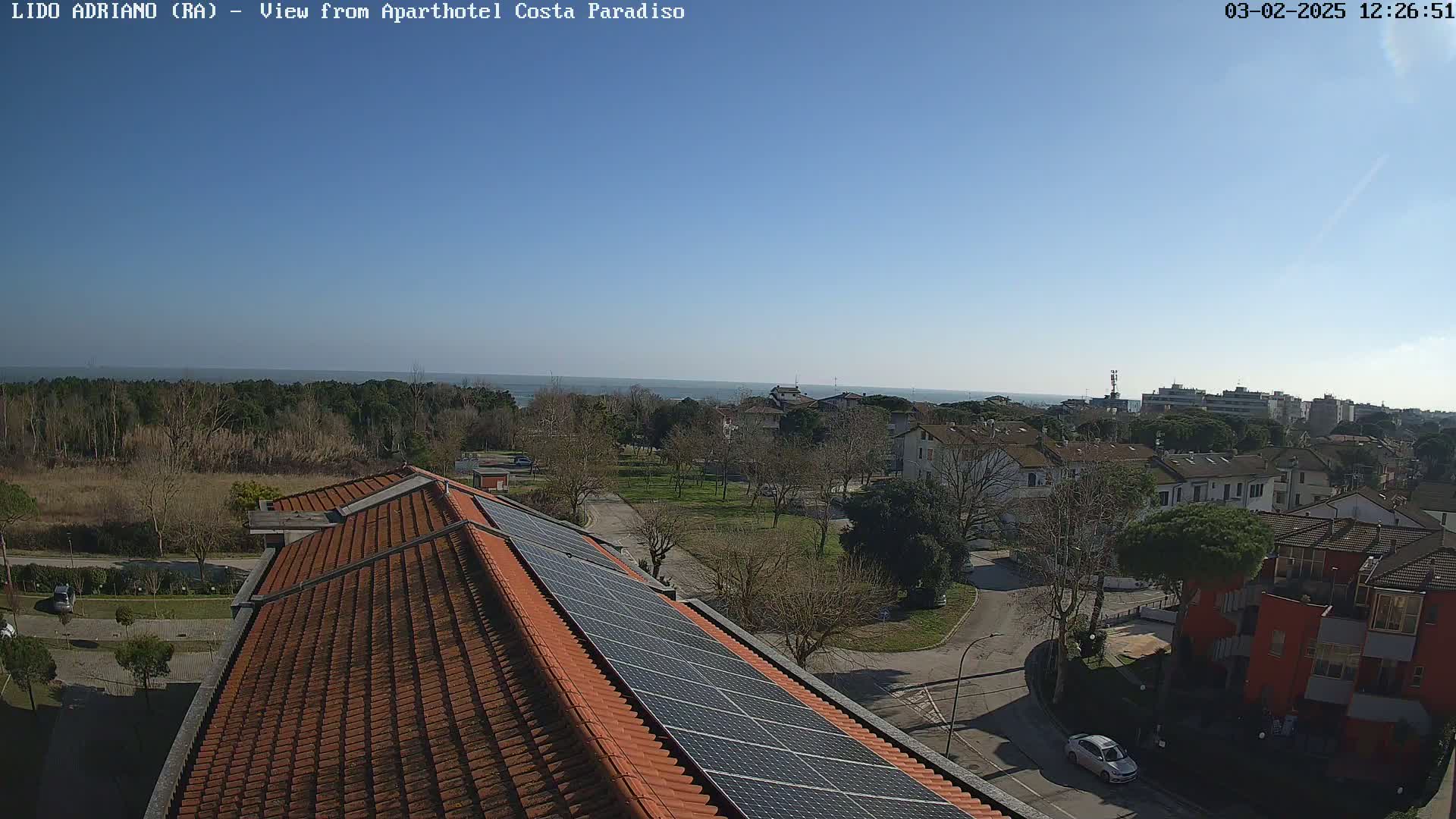 A rooftop with solar panels overlooks a residential area and a calm sea under a clear blue sky.