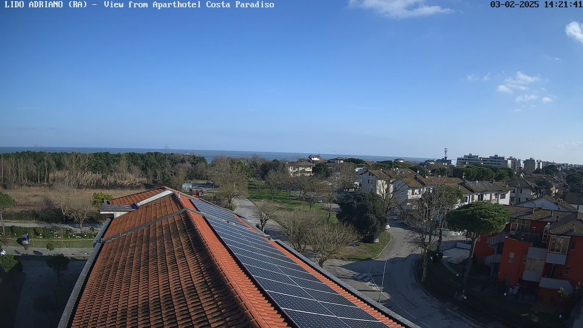 A rooftop with solar panels overlooks a residential area and a calm ocean under a mostly clear blue sky.