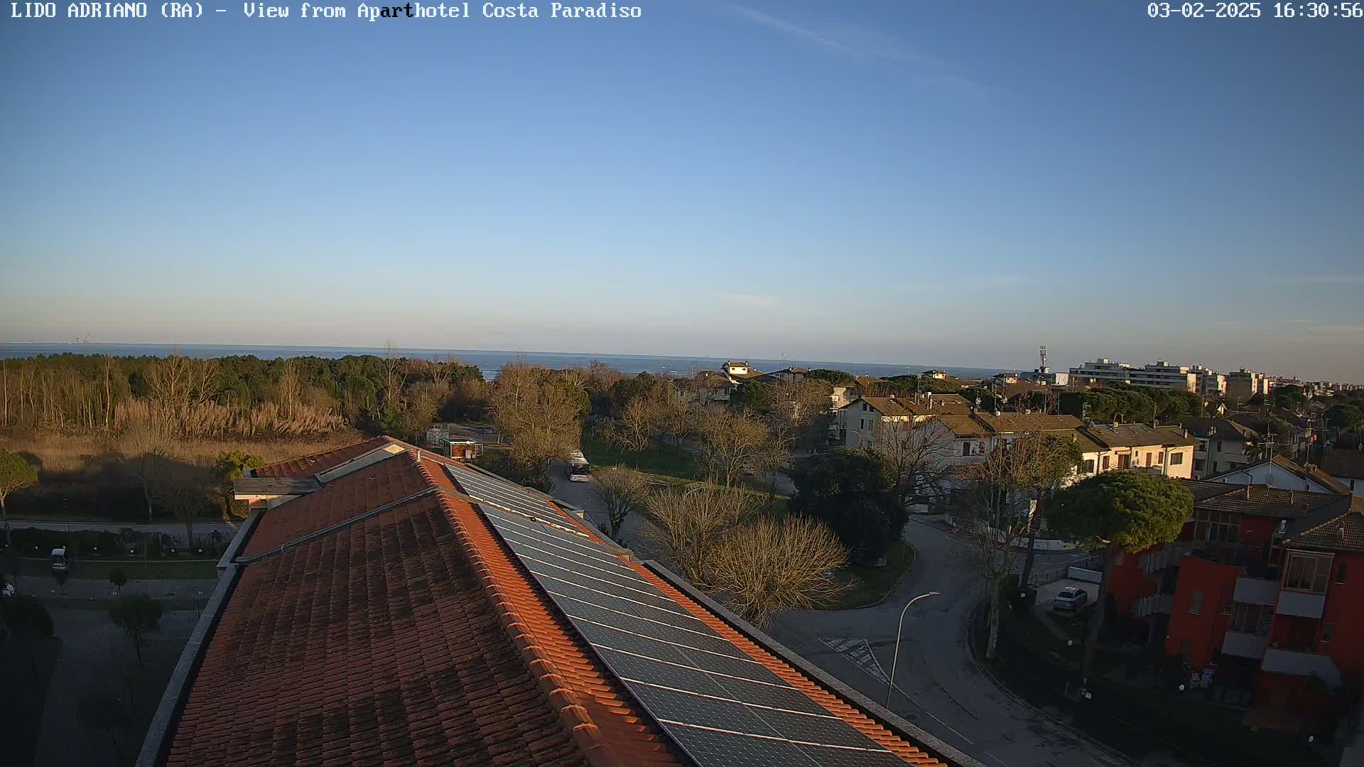 A clear, sunny day overlooks a coastal town with a tiled roof and solar panels in the foreground, and a calm sea in the distance.