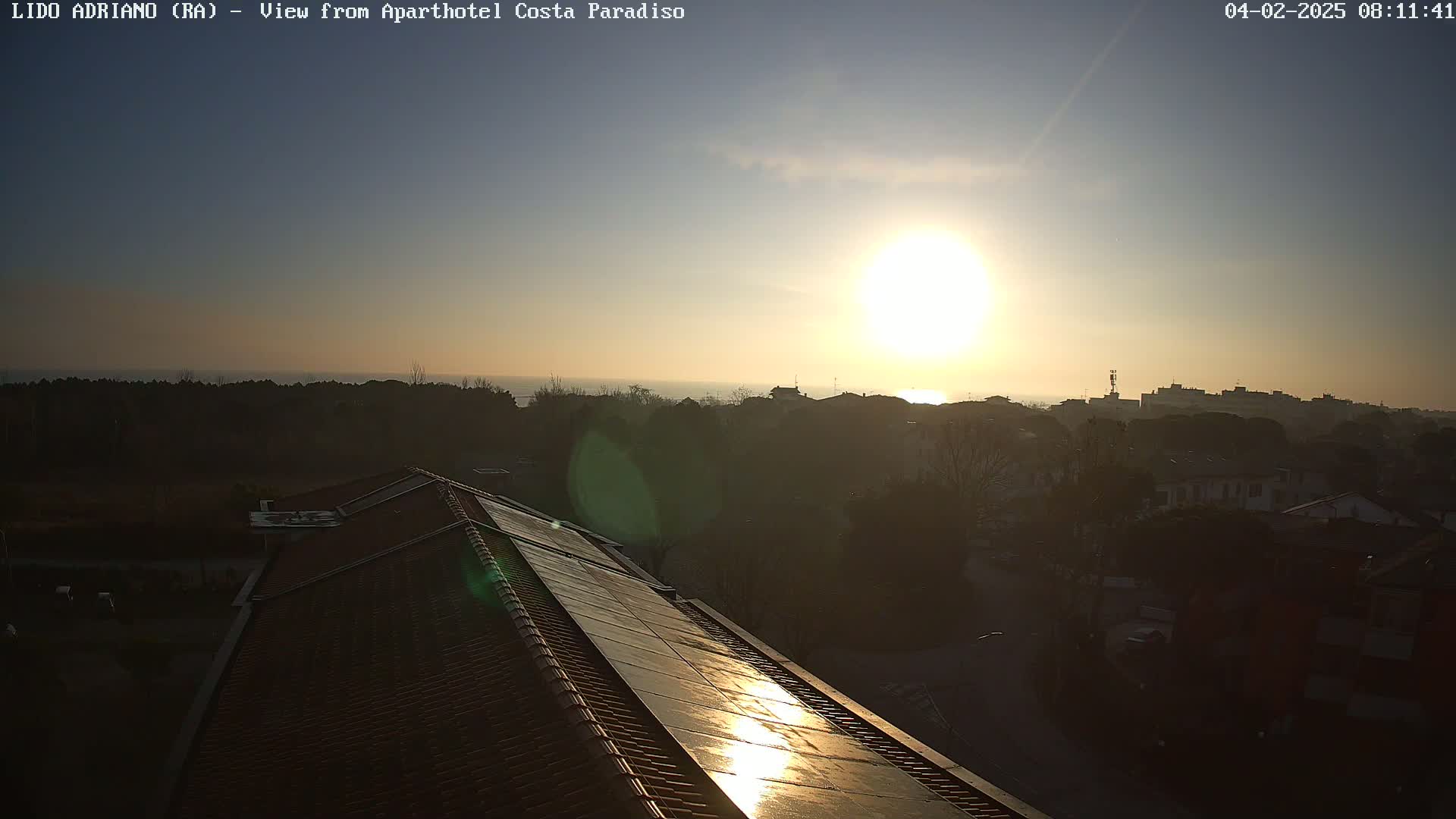 A sunlit view from a rooftop shows a coastal town at sunset, with a calm sea in the distance.
