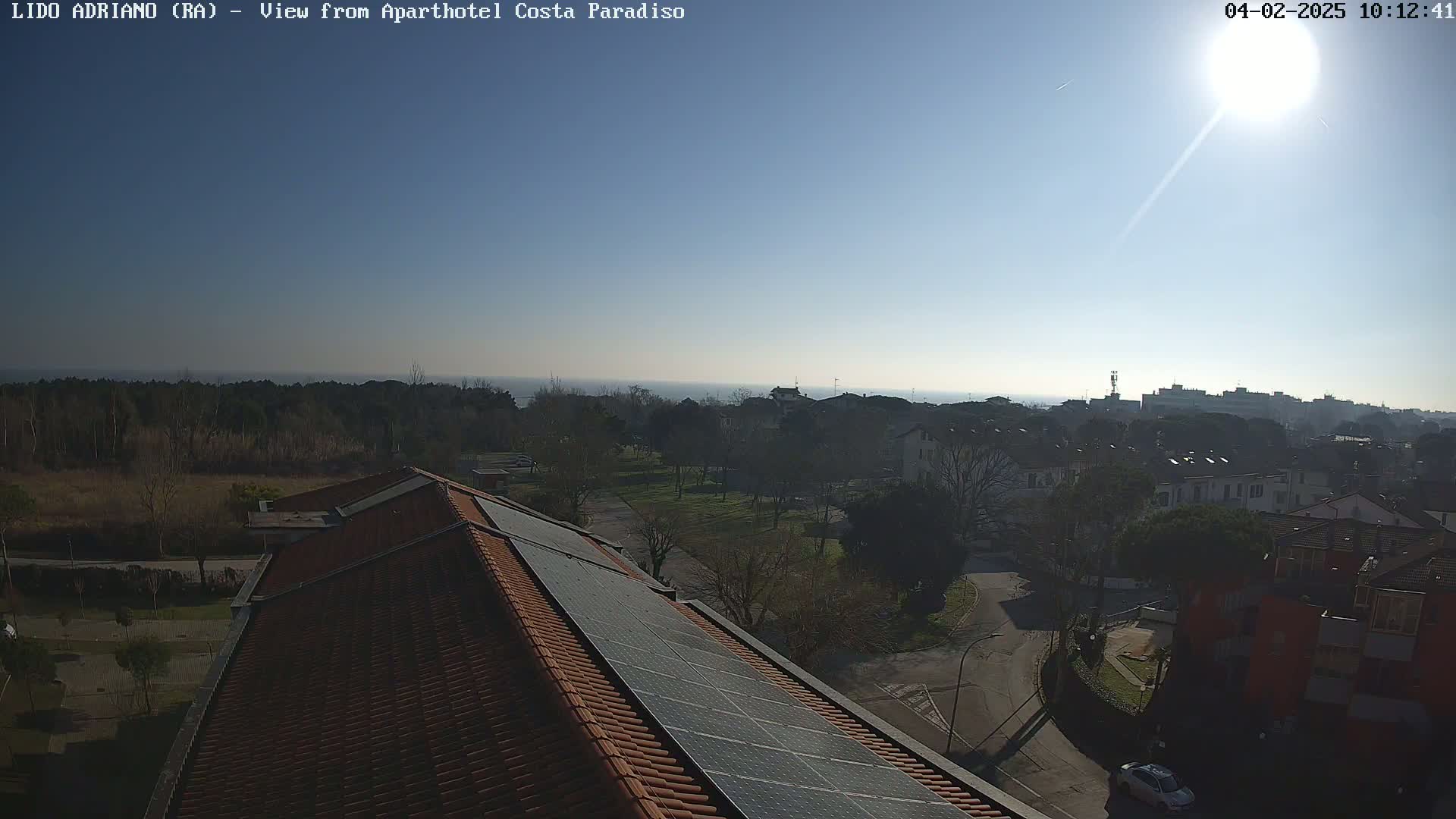 A sunny day overlooking a residential area with a view of the sea in the distance, featuring a building with solar panels on its roof.
