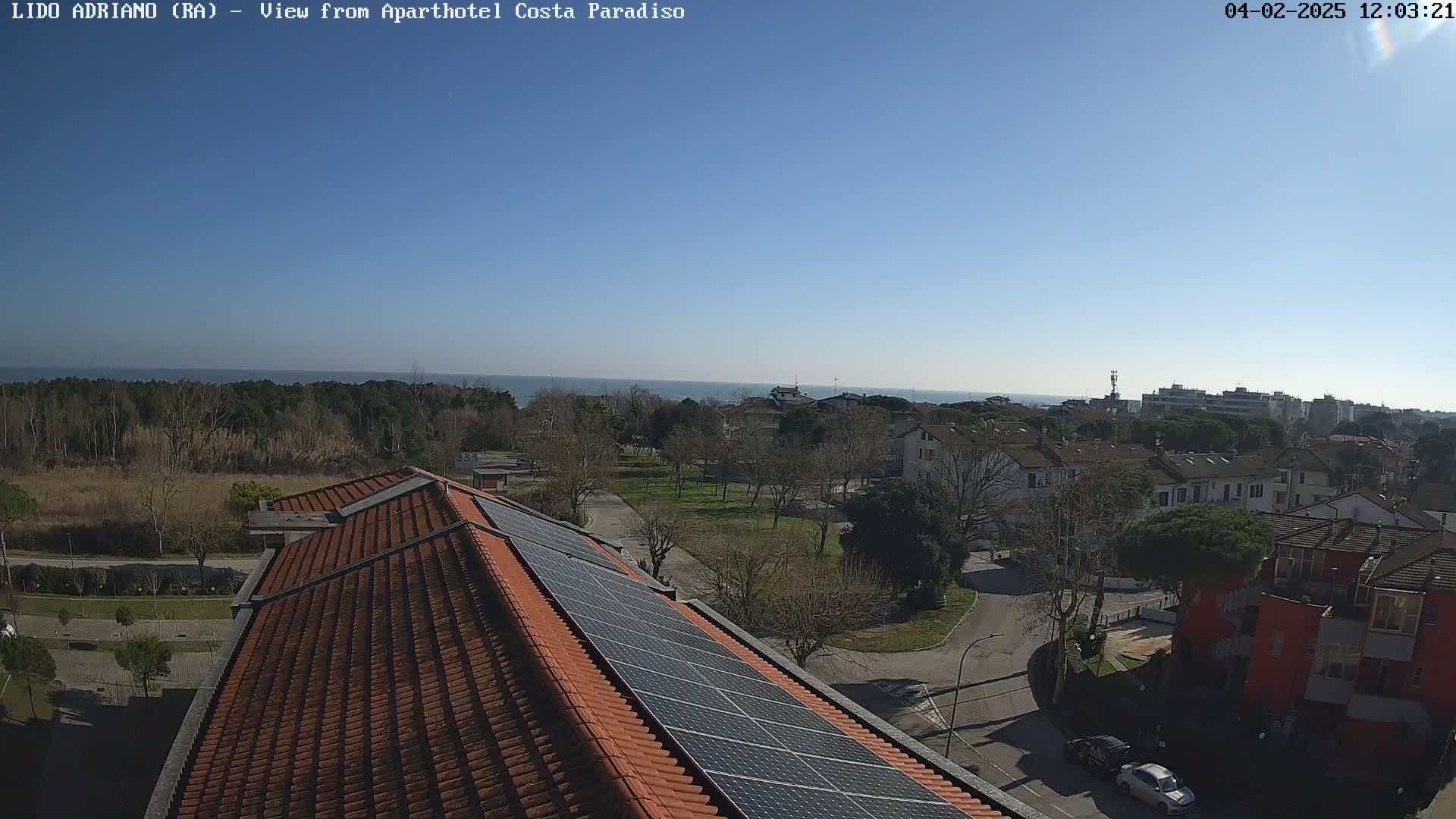A sunny day overlooks a residential area with a tiled roof featuring solar panels, a park, and a calm ocean in the distance.
