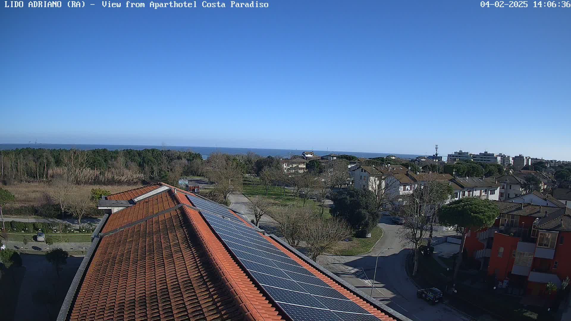 A clear, sunny day reveals a coastal town viewed from a rooftop with solar panels, showing a residential area with trees and a calm ocean in the distance.