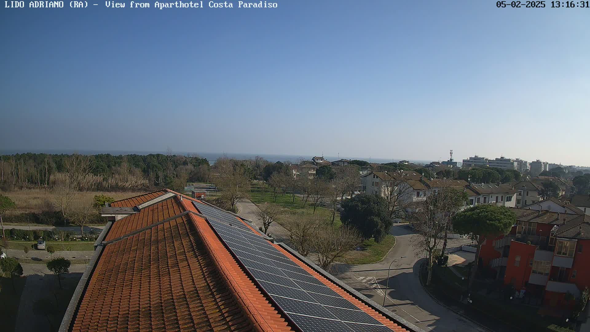 A clear, sunny day view from a rooftop with solar panels overlooking a residential area, a line of trees, and a calm sea in the distance.
