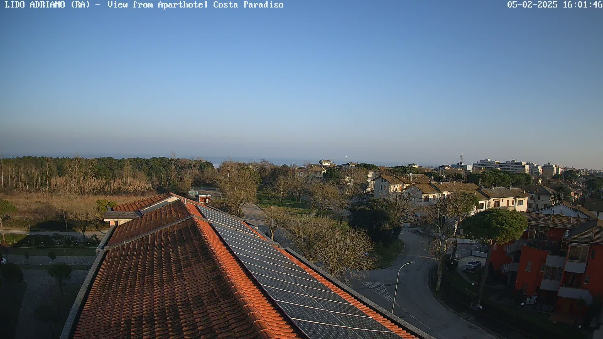 A clear, sunny day reveals a view of a residential area with a tiled roof featuring solar panels, overlooking a tree line and a distant body of water.