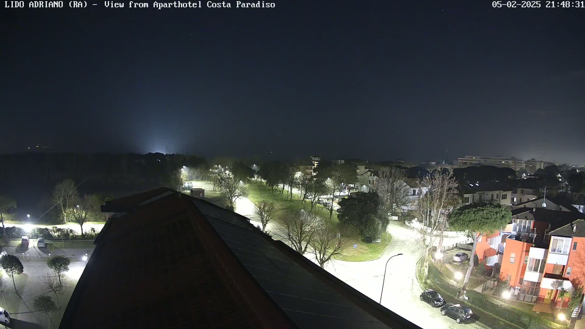 A nighttime view of a residential area with well-lit streets and parked cars under a dark, slightly hazy sky.