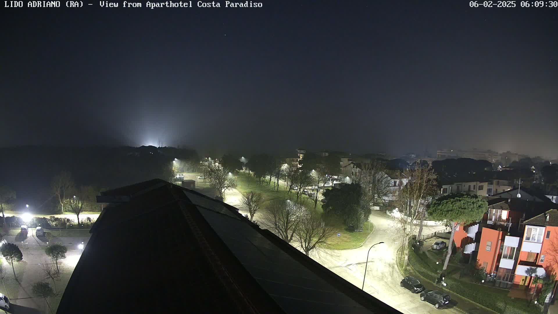 A nighttime, foggy view from a rooftop shows a residential area with streetlights illuminating roads and buildings.