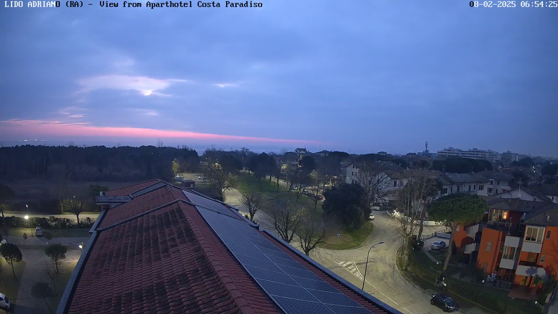 A twilight view from a rooftop with solar panels overlooks a residential area and a calm sea under a mostly clear sky.