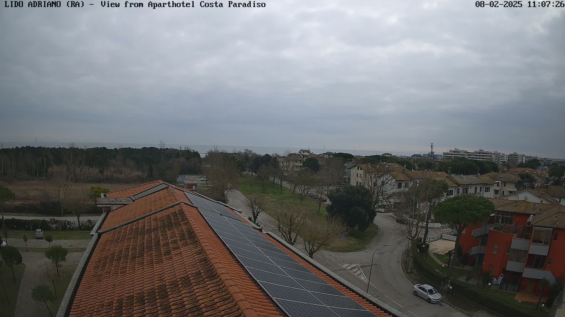 A view from a rooftop with solar panels shows a residential area near a calm sea under an overcast sky.