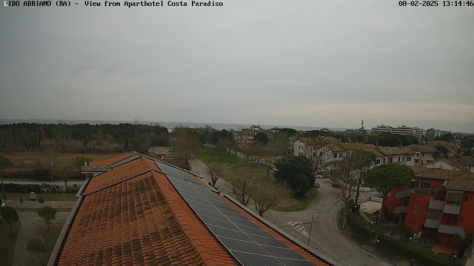 An overcast day reveals a coastal town viewed from a building rooftop with solar panels, featuring residential buildings, streets, and a calm sea in the distance.