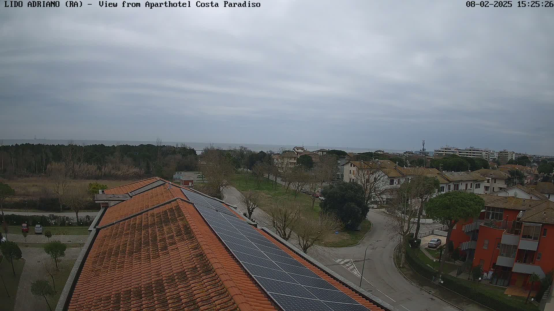 An overhead view of a residential area with a tiled roof and solar panels, overlooking a calm sea under a cloudy sky.