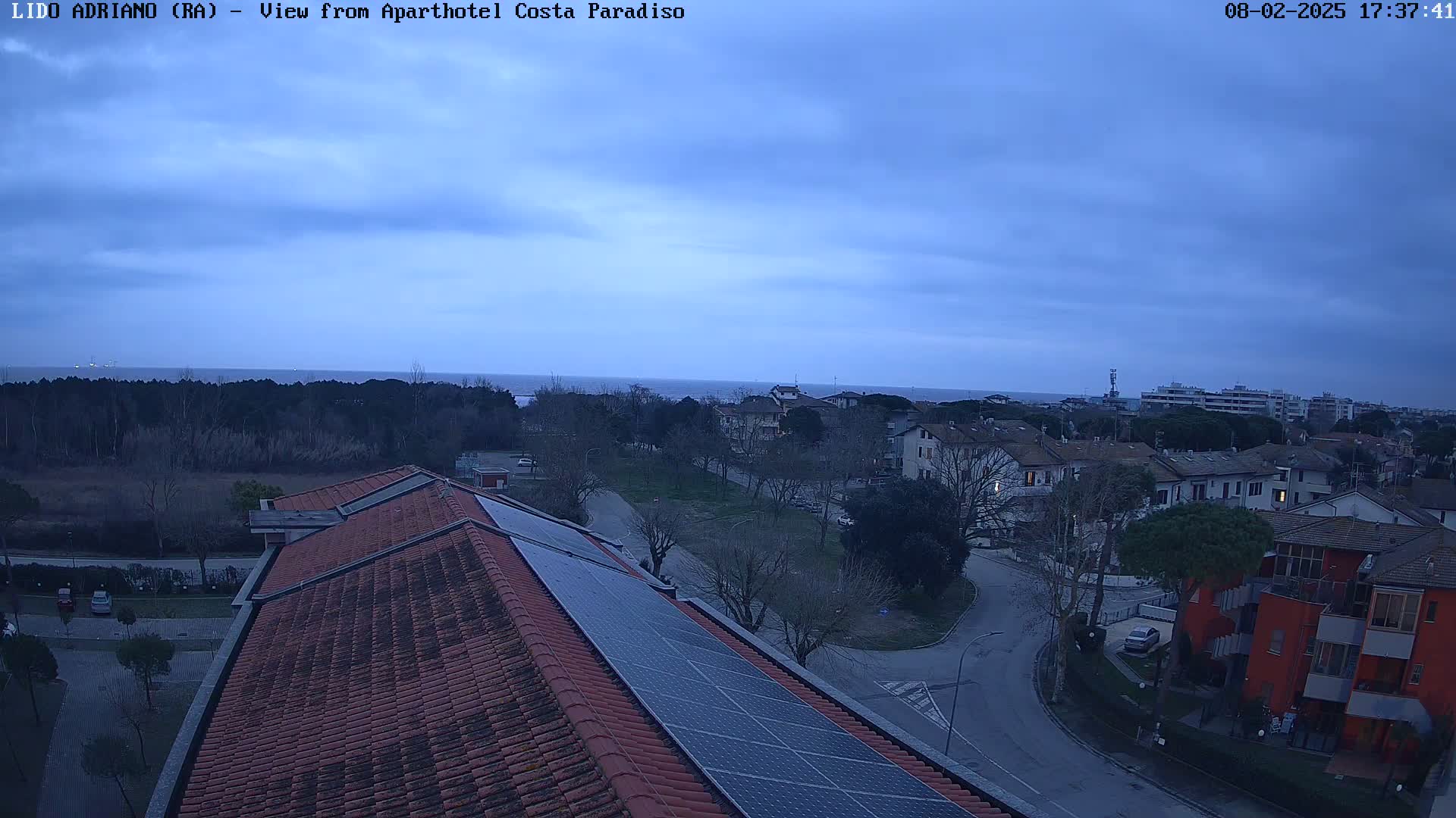 A view from a rooftop with solar panels shows a residential area near a calm sea under a cloudy, dusky sky.