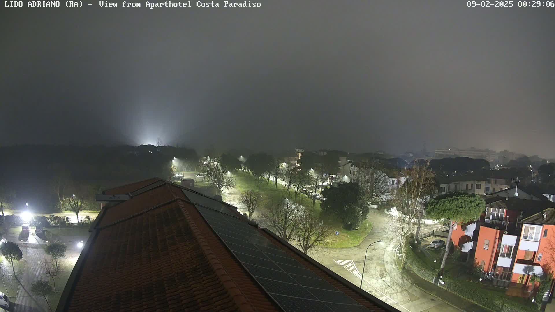 A nighttime view of a residential area shrouded in fog, showing rain-slicked streets and buildings illuminated by streetlights.