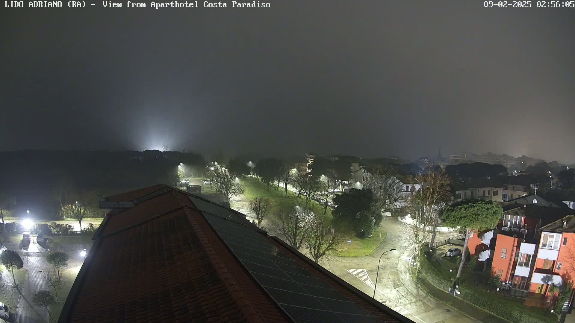 A nighttime, foggy view of a residential area with streetlights illuminating wet roads and buildings, seen from a rooftop with solar panels.