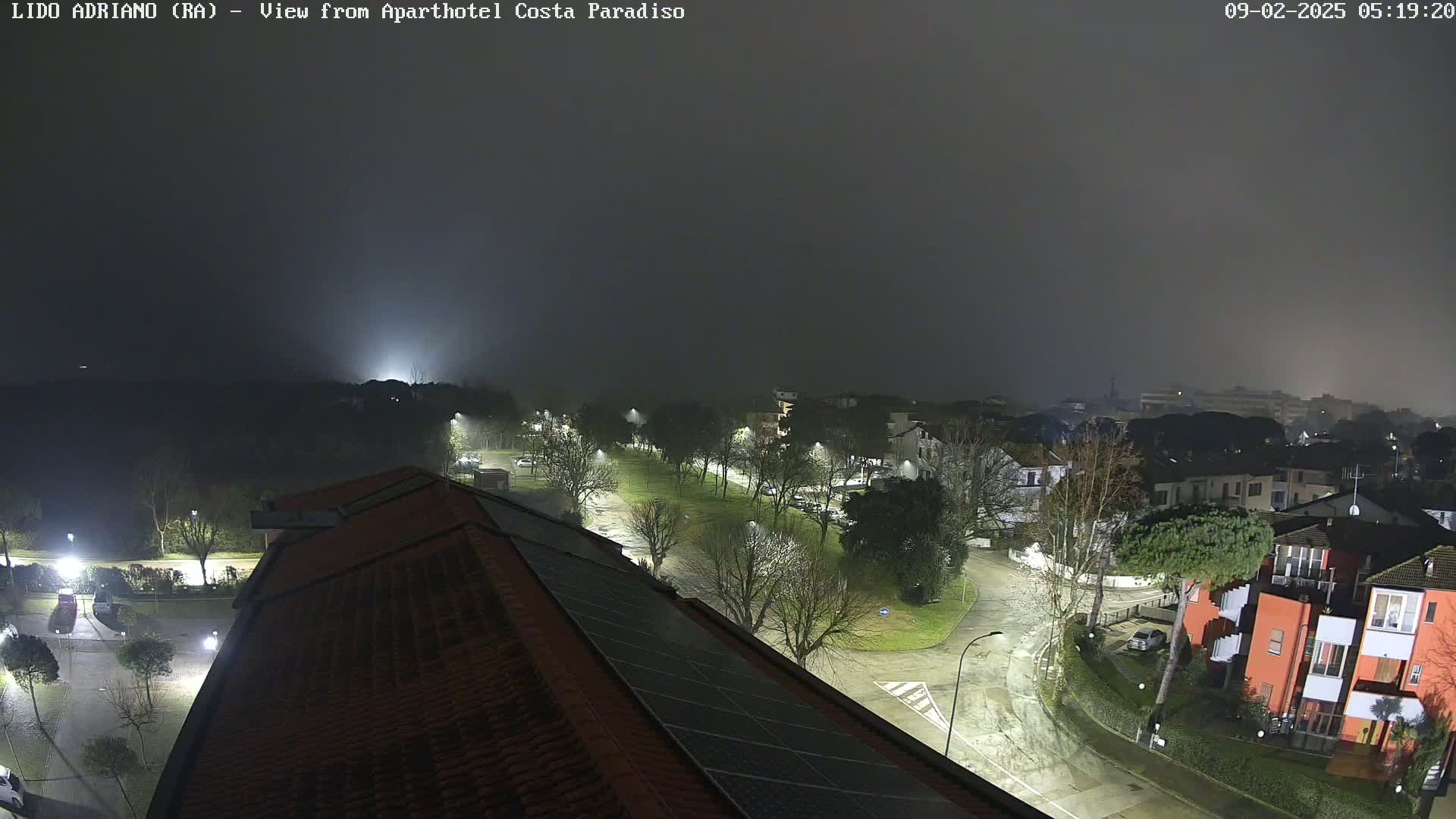 A nighttime view of a residential area with fog, showing tree-lined streets, buildings, and a rooftop with solar panels.