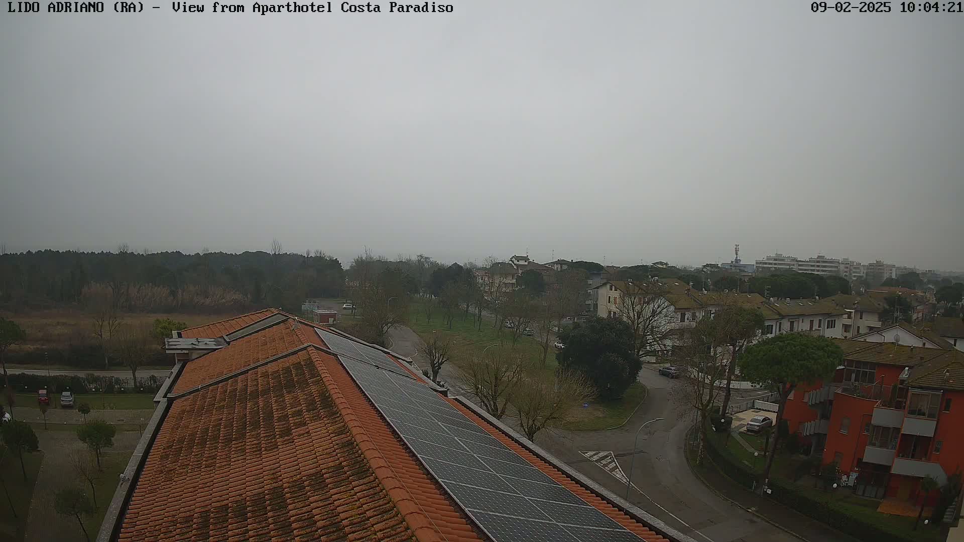 A view of a residential area on an overcast day, showing a building with solar panels on its roof and surrounding houses and trees.