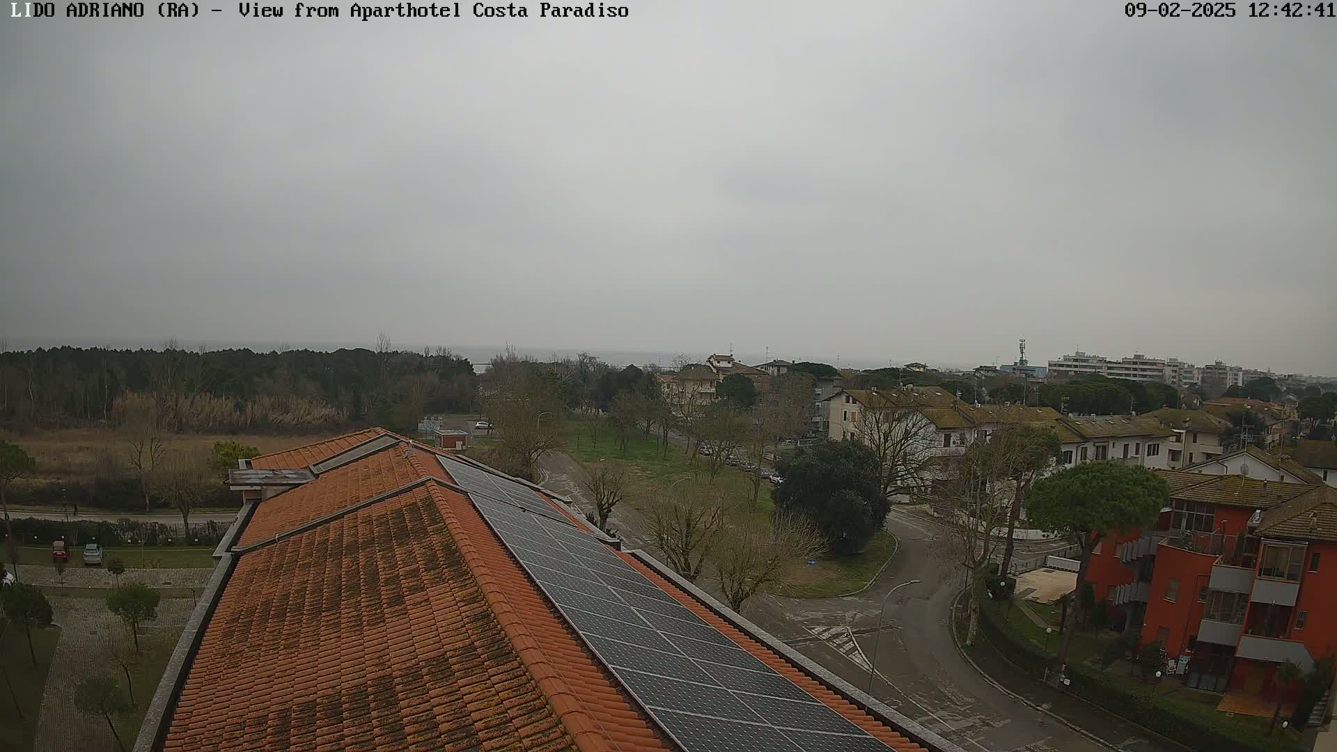 A view of a town on an overcast day, seen from a rooftop with solar panels, shows a residential area with a distant glimpse of the sea.