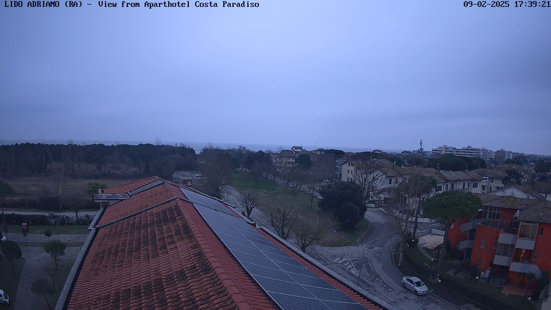 A rooftop with solar panels overlooks a residential area near a calm sea under an overcast sky.