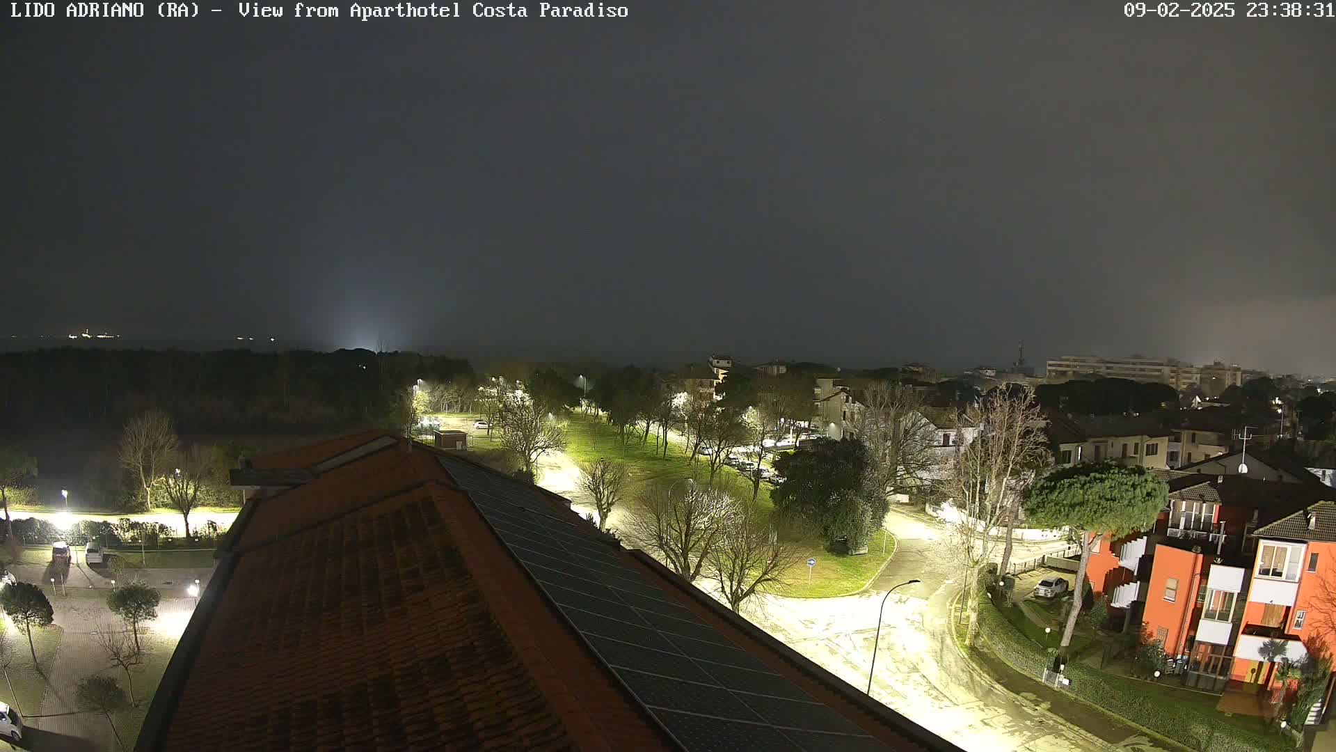 A nighttime view of a residential area with lightly snow-covered roads and trees, under a dark, cloudy sky.