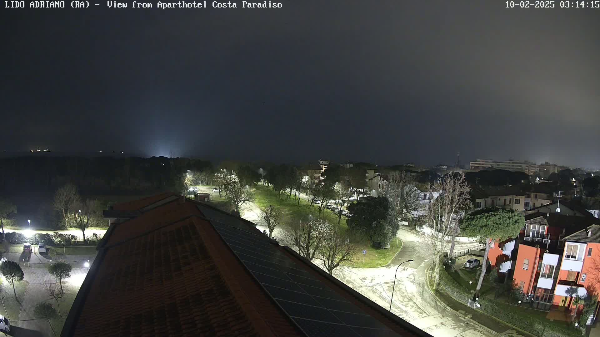 A nighttime, foggy view of a residential area with streetlights illuminating roads and buildings, some of which have solar panels on their roofs.