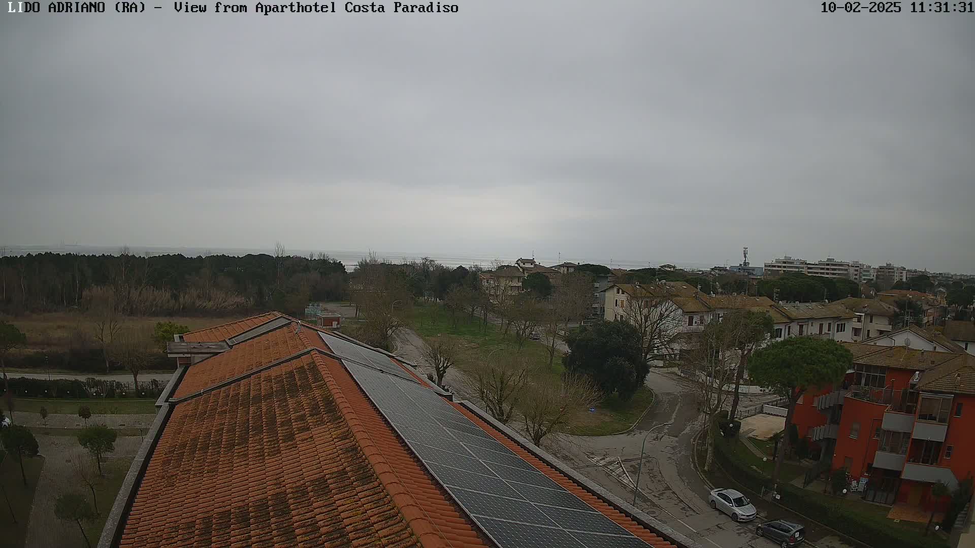 A view from a rooftop with solar panels shows a cloudy day overlooking a residential area near a calm body of water and a line of trees.