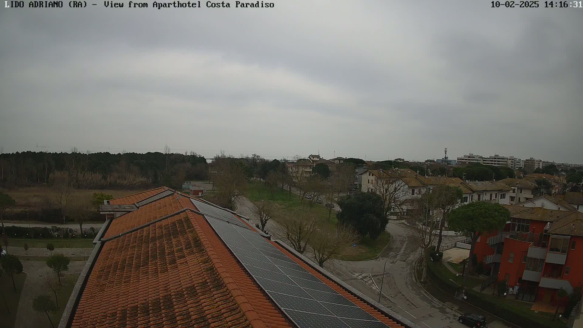 An overcast day reveals a view of a residential area with a tiled rooftop equipped with solar panels, overlooking a park-like space and the sea in the distance.