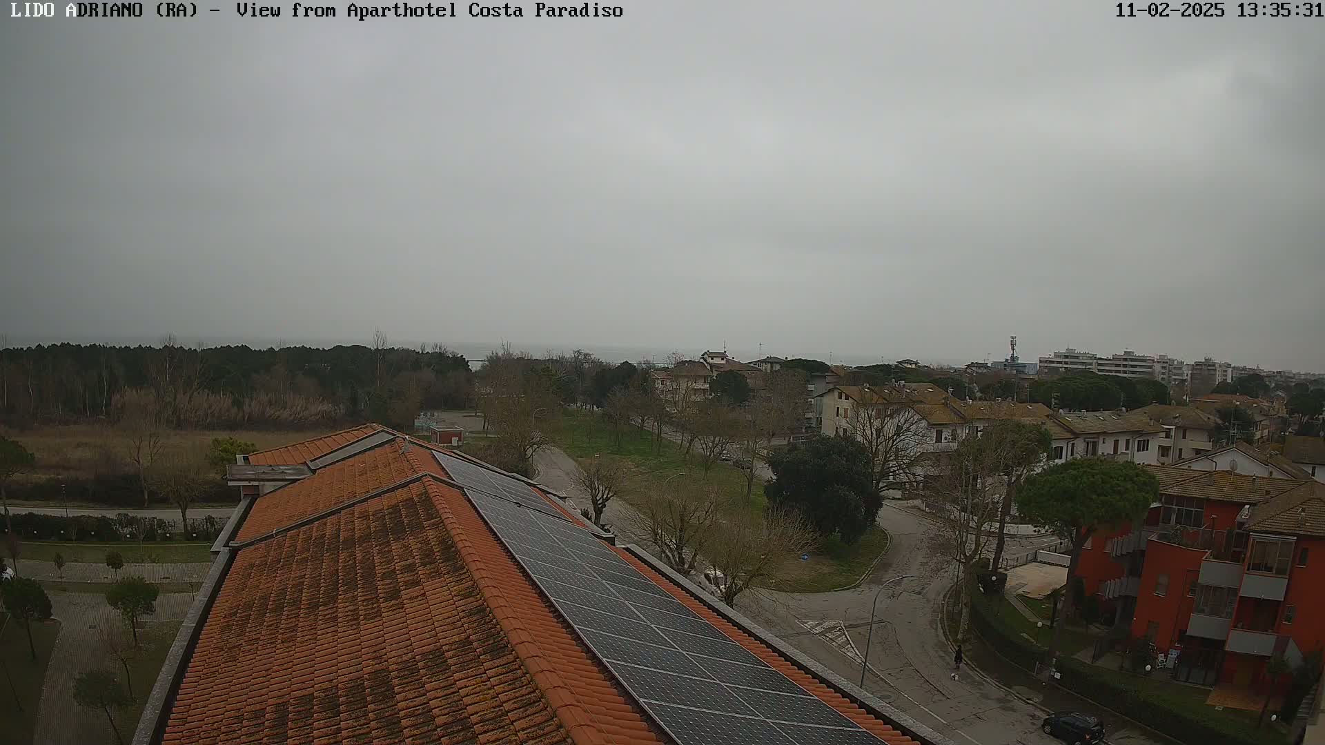 A view from a rooftop with orange tiles and solar panels shows a residential area and a body of water under an overcast sky.