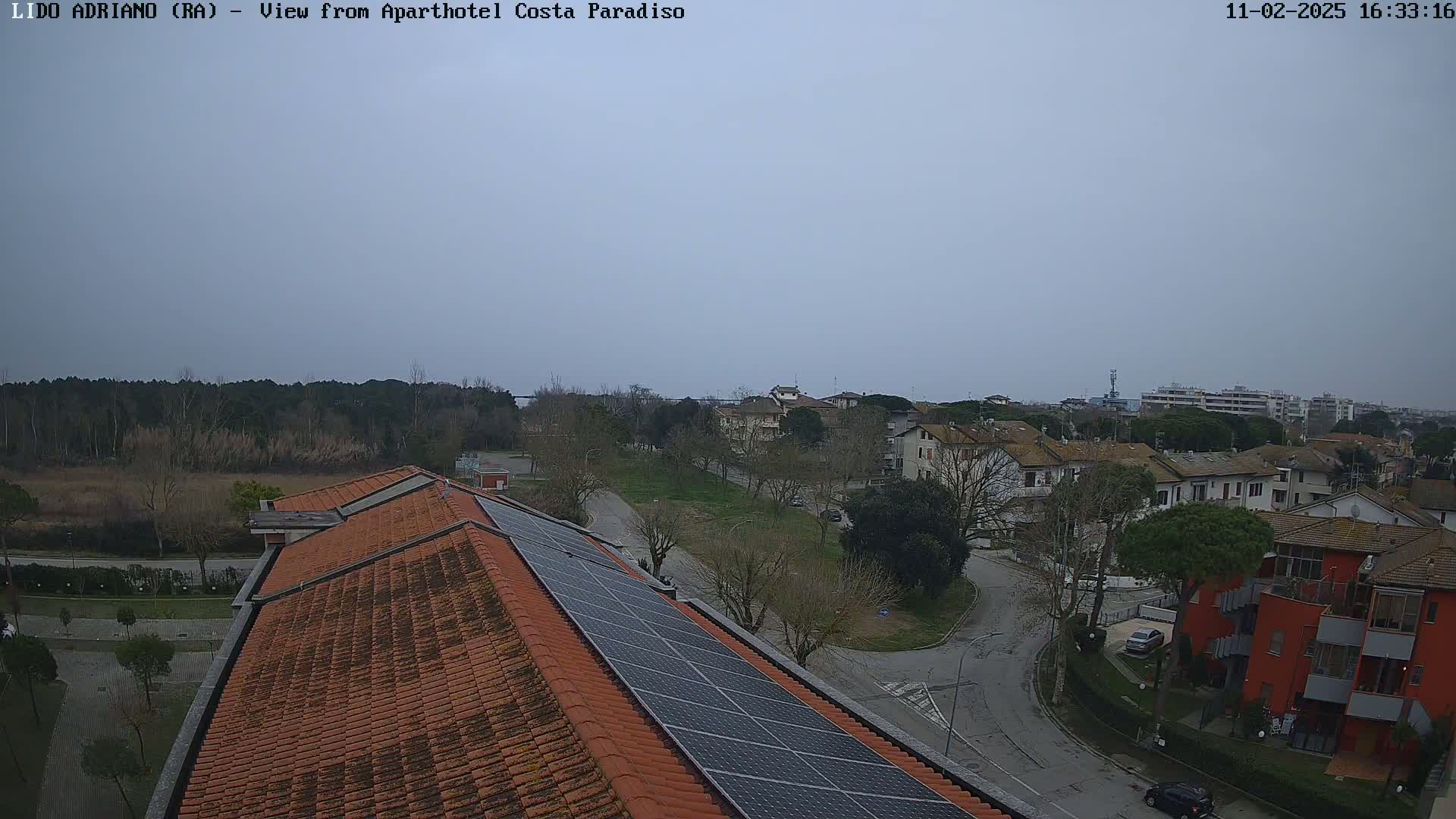 An overcast day reveals a rooftop with solar panels overlooking a residential area with low buildings and sparse trees.
