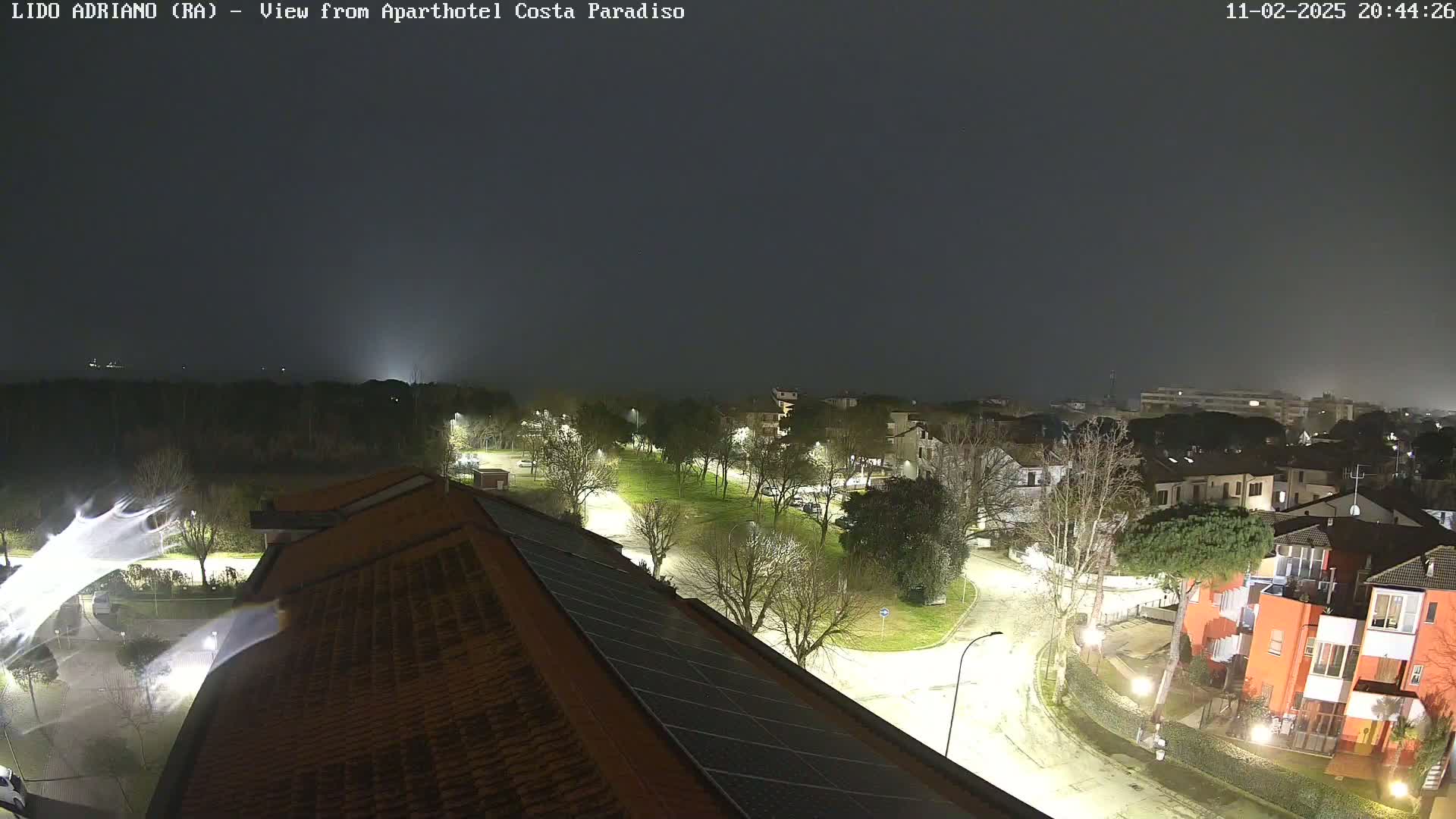 A nighttime view of a residential area with a park, illuminated streets, and a partially visible body of water in the distance under a dark, cloudy sky.