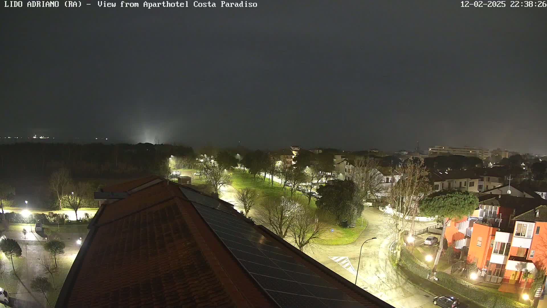 A nighttime, rain-slicked view of a residential area with streetlights illuminating roads and buildings, and a dark, overcast sky.