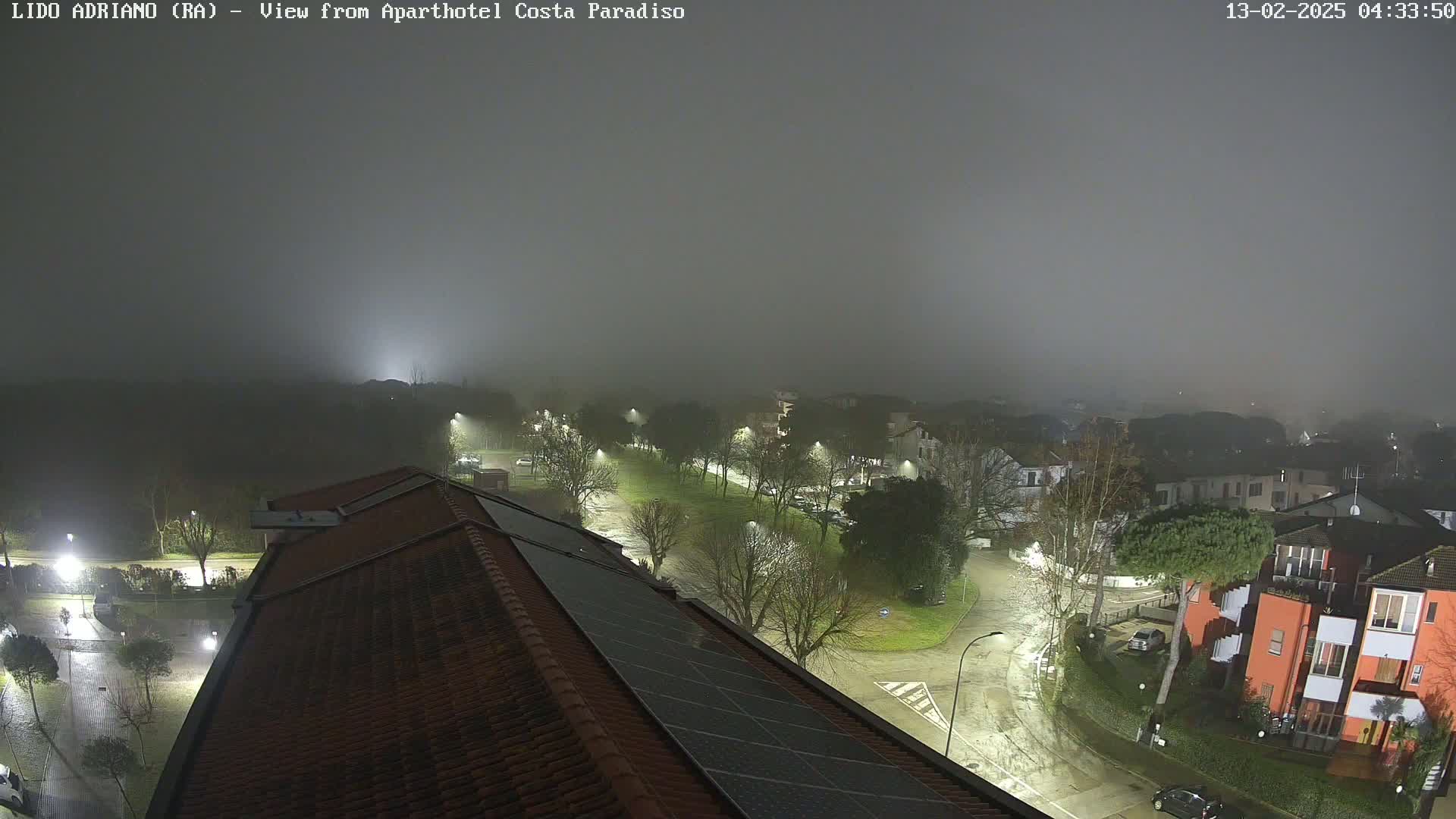 A nighttime view of a residential area shrouded in fog, showing a street with parked cars and buildings, illuminated by streetlights, as seen from a rooftop with solar panels.