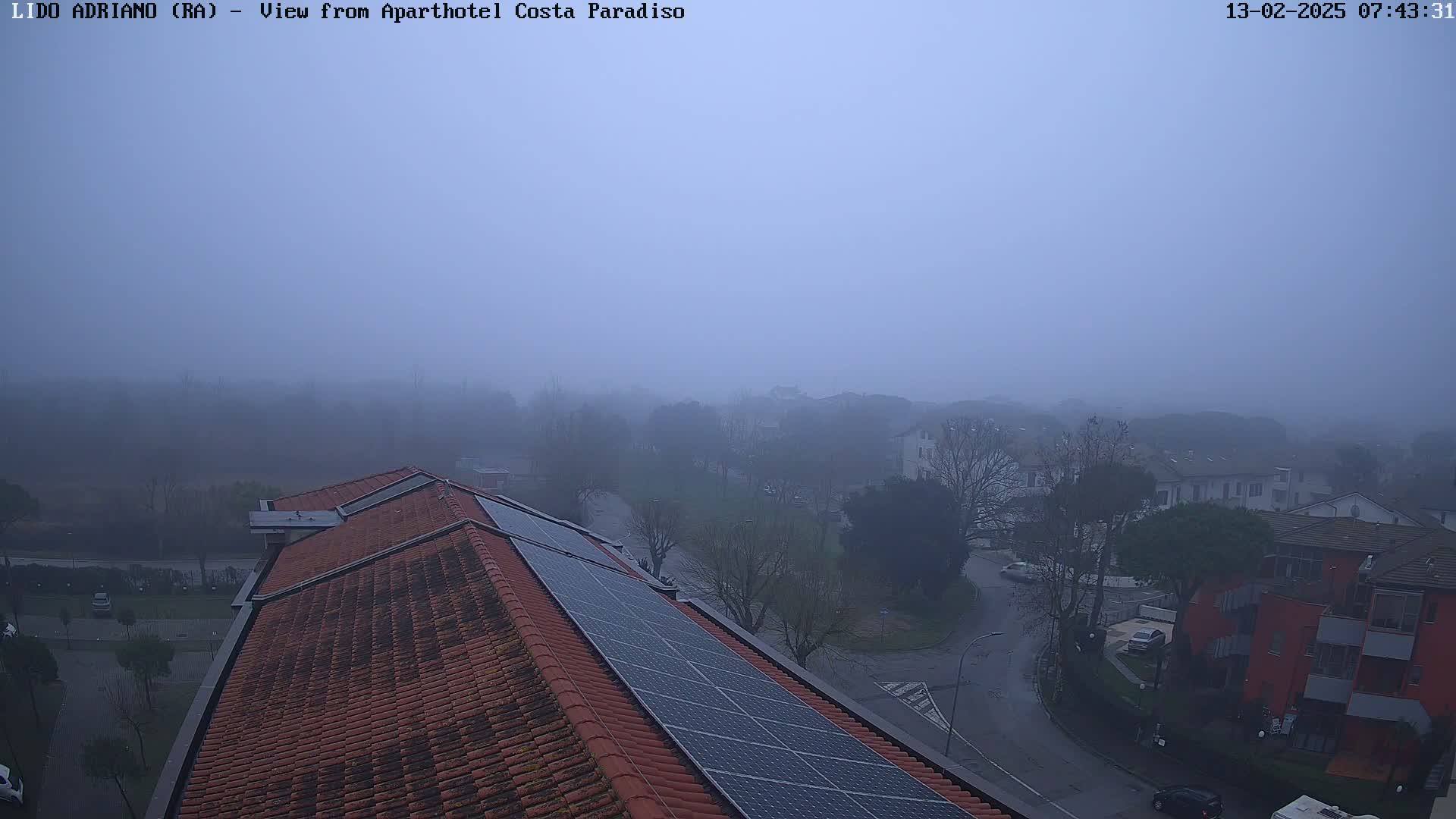 A foggy, overcast view from a rooftop shows a residential area with red-tiled roofs and solar panels, a street, and several trees.