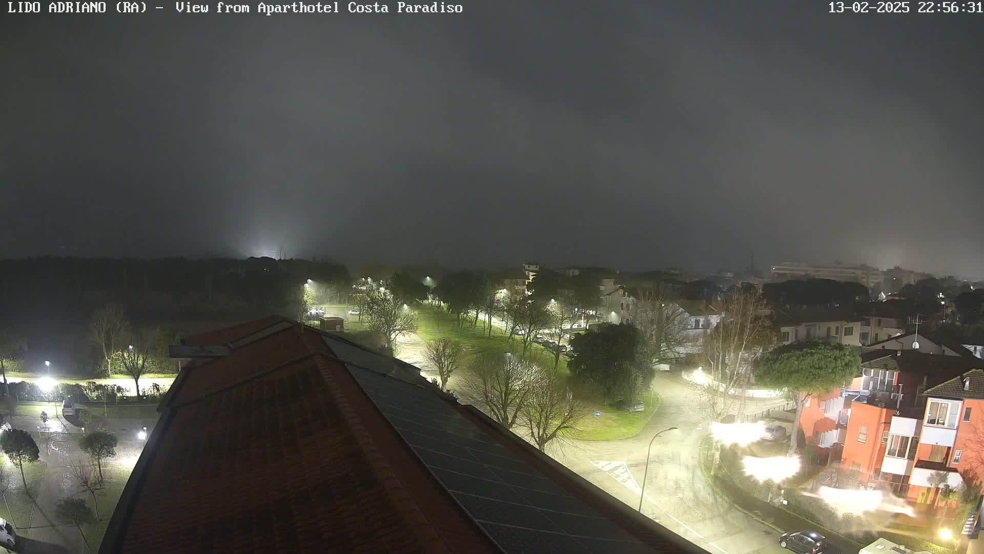 A nighttime, foggy view of a residential area with illuminated streets and buildings, seen from a rooftop with solar panels.