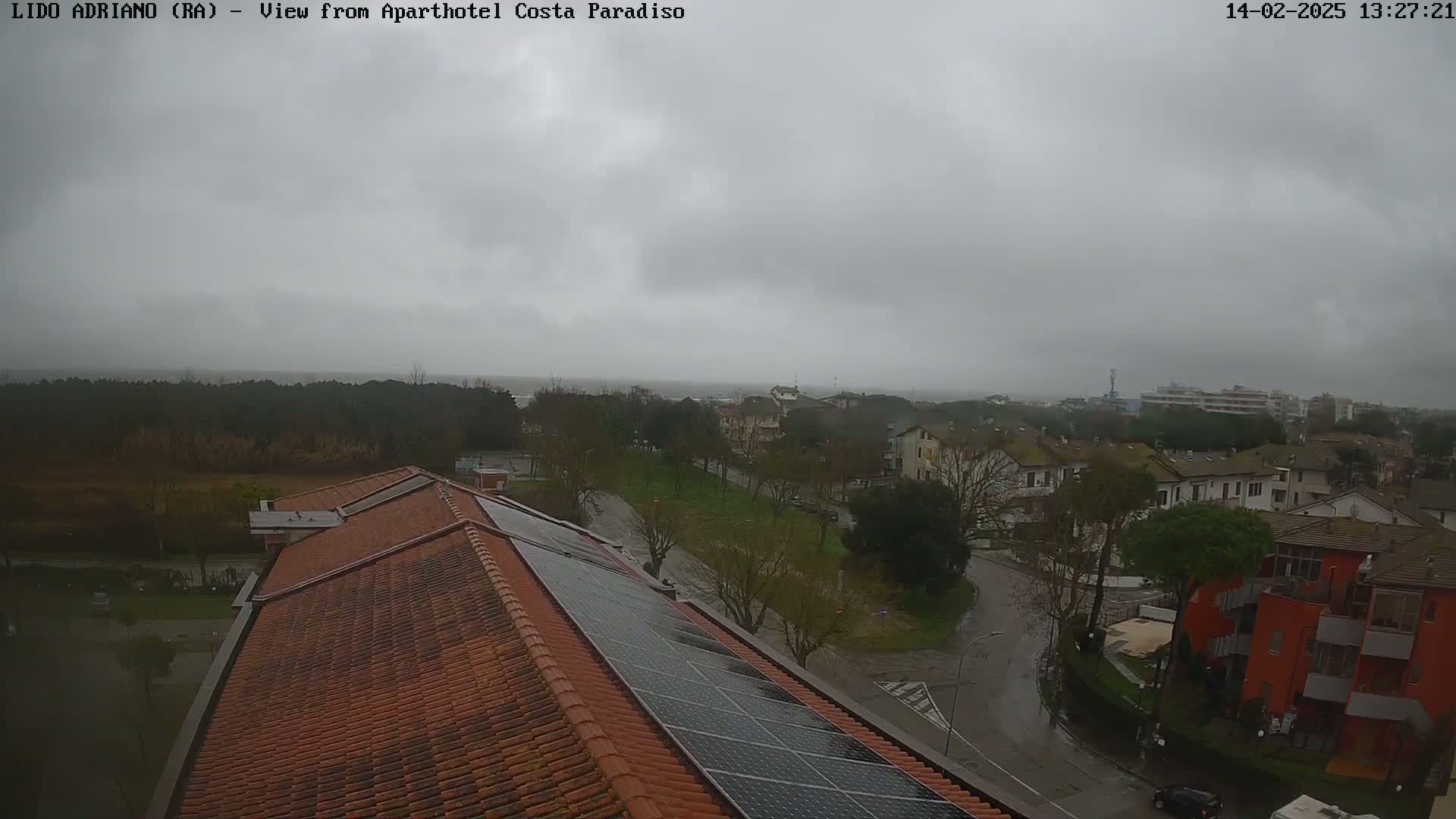 A view from a rooftop with solar panels shows a residential area on a cloudy, wet day, with a body of water visible in the distance.